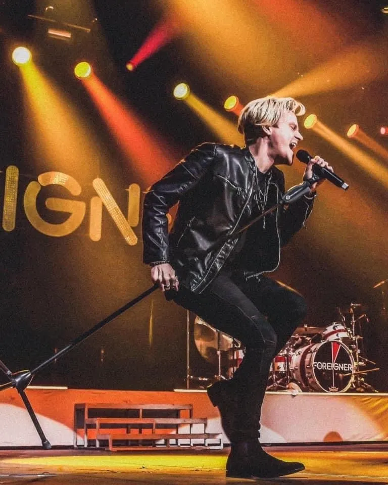 Geordie brown holds a mic stand and bends forward dramatically with bright stage lights behind him.